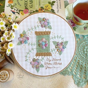 May include: A circular embroidery hoop featuring a light green thread spool centered among floral fabric appliqués on a white and beige striped fabric. The text "Joy Blooms Where Stitches Grow" is embroidered in brown thread below the spool.