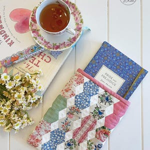 May include: A patchwork book sleeve with a pink border, holding a copy of "Pride and Prejudice." The sleeve features a geometric design with floral and bird patterns. A teacup and saucer with a floral design sit atop books, with a bouquet of white flowers.