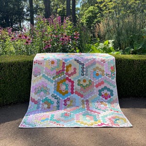 Puede incluir: Una colcha colorida con un patrón hexagonal en tonos de rosa, verde, amarillo y azul. La colcha está hecha de varias telas florales y se muestra sobre una superficie cubierta de hierba.