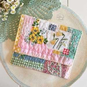 May include: A handcrafted patchwork wallet with floral embroidery and diverse fabric patterns. The wallet incorporates pink, yellow, blue, and green fabrics, a scalloped edge, and a gingham pattern.