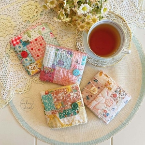 May include: A collection of four quilted fabric coasters in various floral and patchwork patterns. Each coaster features a unique combination of colors and textures, with button closures. A cup of tea and flowers are also in the image.
