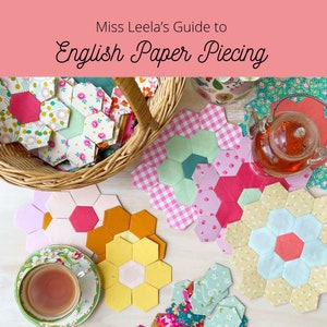May include: A close-up of a table with several colourful fabric hexagons arranged in a flower shape. The hexagons are made of different fabrics and colours, including pink, green, yellow, and orange. The hexagons are arranged in a circle, with a smaller hexagon in the centre. The hexagons are surrounded by a white surface. The text "Miss Leela's Guide to English Paper Piecing" is written in black text on a pink background at the top of the image.