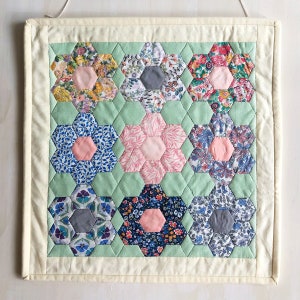 May include: A white fabric wall hanging with a green background and a patchwork design of nine hexagonal floral patterns in shades of pink, blue, and grey.