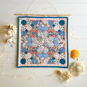 May include: A teal framed wall hanging with a pink and blue patchwork quilt design. The quilt is made up of small squares of fabric in various patterns. The quilt is hung on a white wall with string lights.