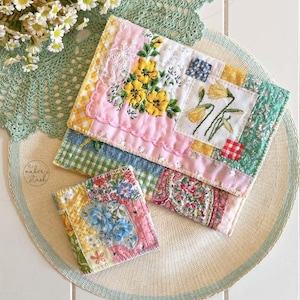 May include: A collection of handmade fabric items, including a small square coaster and two larger pouches. The items feature patchwork designs with floral embroidery and various patterned fabrics in pink, yellow, blue, and green.
