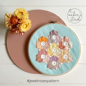 May include: A light blue embroidery hoop with a colorful hexagonal quilt pattern in shades of pink, orange, yellow, and purple. The hoop is on a pink surface with the text "#jewelrosespattern" below it.