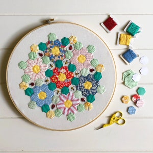 以下が含まれることがあります： 六角形のデザインのカラフルなパッチワークキルトブロック。ブロックは赤、黄、緑、青、白の様々な生地で作られています。ブロックは白い背景で囲まれています。