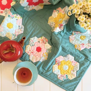 以下が含まれることがあります： カラフルな花柄の入ったパッチワークデザインのティールブルーのキルト。キルトは中央に花柄のモチーフが入った六角形の形をしています。キルトは白い表面にティーポットとティーカップと一緒に置かれています。