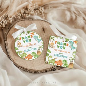Può includere: Due etichette regalo a tema dinosauri con nastro bianco. Una è rotonda, l'altra quadrata. Entrambe presentano il testo "THANK YOU FOR STOMPING BY Love, SEBASTIAN" con illustrazioni di dinosauri, palme e un vulcano.