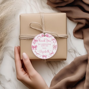 May include: A gift box wrapped in tan paper, tied with twine, and a round tag. The tag features a pink bow design and the text "Thank You for celebrating with me, Love, Elizabeth."