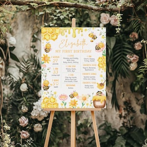 May include: A decorative sign on an easel for a first birthday celebration. The sign features a bee theme with the name "Elizabeth" and details like weight, height (33 inches), and favorite things. The design includes bees, flowers, and honeycomb.