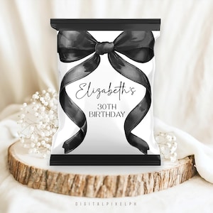 Könnte beinhalten: Eine weiße Party-Geschenktüte mit einem schwarzen Schleifen- und Banddesign. Der Text "Elizabeth's 30th Birthday" ist in eleganter Schrift dargestellt. Die Tüte steht auf einer Holzscheibe mit weißen Blumen.
