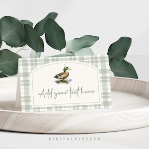 May include: A folded place card with a watercolor illustration of a duck wearing a party hat. The card has a light green plaid pattern and the text "Add your text here". The card is on a white wooden tray with green foliage in the background.