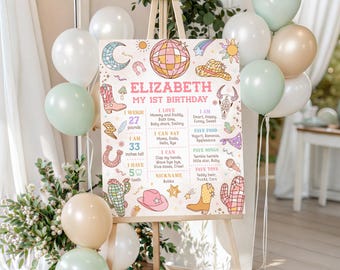 May include: A rectangular birthday milestone sign featuring the text Elizabeth My 1st Birthday, surrounded by colorful illustrations of a disco ball, cowboy boots, cacti, and stars. The board lists personal details including 27 pounds and 33 inches tall.