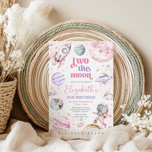 May include: A pink and purple invitation with a space theme. The text reads "Two the Moon" and "Elizabeth's 2nd Birthday." The design includes a rocket ship, planets, a UFO, and an astronaut. The invitation is set against a woven background.