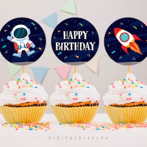 May include: Three cupcakes with white frosting and colorful sprinkles. Each cupcake has a round topper with a space theme. One shows an astronaut, another says "Happy Birthday," and the last has a rocket.