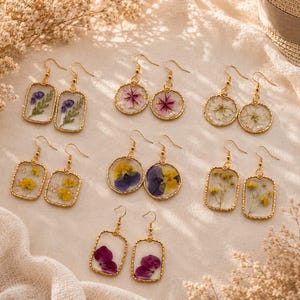 Puede incluir: Siete pares de pendientes colgantes con flores prensadas en resina transparente y marcos de metal dorado. La colección incluye formas rectangulares y circulares con especímenes florales en tonos morados, rosas, blancos y amarillos.