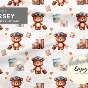 May include: A seamless pattern featuring a brown teddy bear wearing aviator goggles and carrying a suitcase. The pattern also includes hearts, cameras, and luggage tags. The text "EXTENSIBLE JERSEY 95% COTTON 5% ELASTHANNE" is at the top of the image. The text "Exclusive Enjoy Tissus & Mercerie" is at the bottom of the image.
