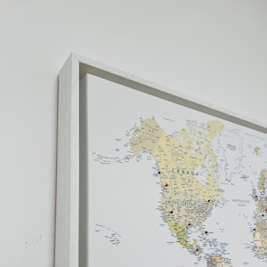 May include: A framed world map with push pins marking locations. The map is in a white frame and features a detailed depiction of the continents and oceans. The map is in a neutral colour palette with blue text and push pins in red and black.