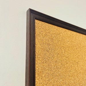 May include: A corkboard with a dark brown wooden frame. The corkboard is made of natural cork and has a textured surface.