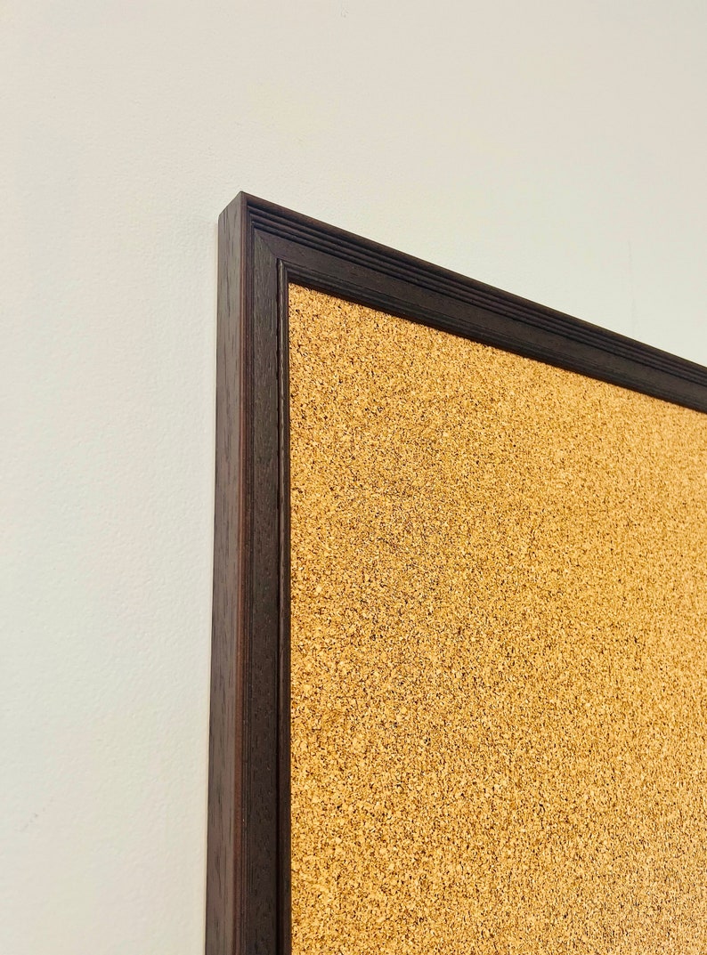 May include: A corkboard with a dark brown wooden frame. The corkboard is made of natural cork and has a textured surface.