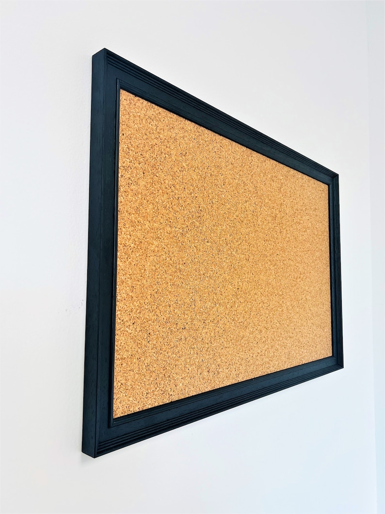 May include: A corkboard with a black frame. The corkboard is mounted on a white wall.