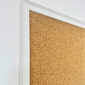 May include: A white framed corkboard with a brown cork surface. The corkboard is mounted on a white wall.