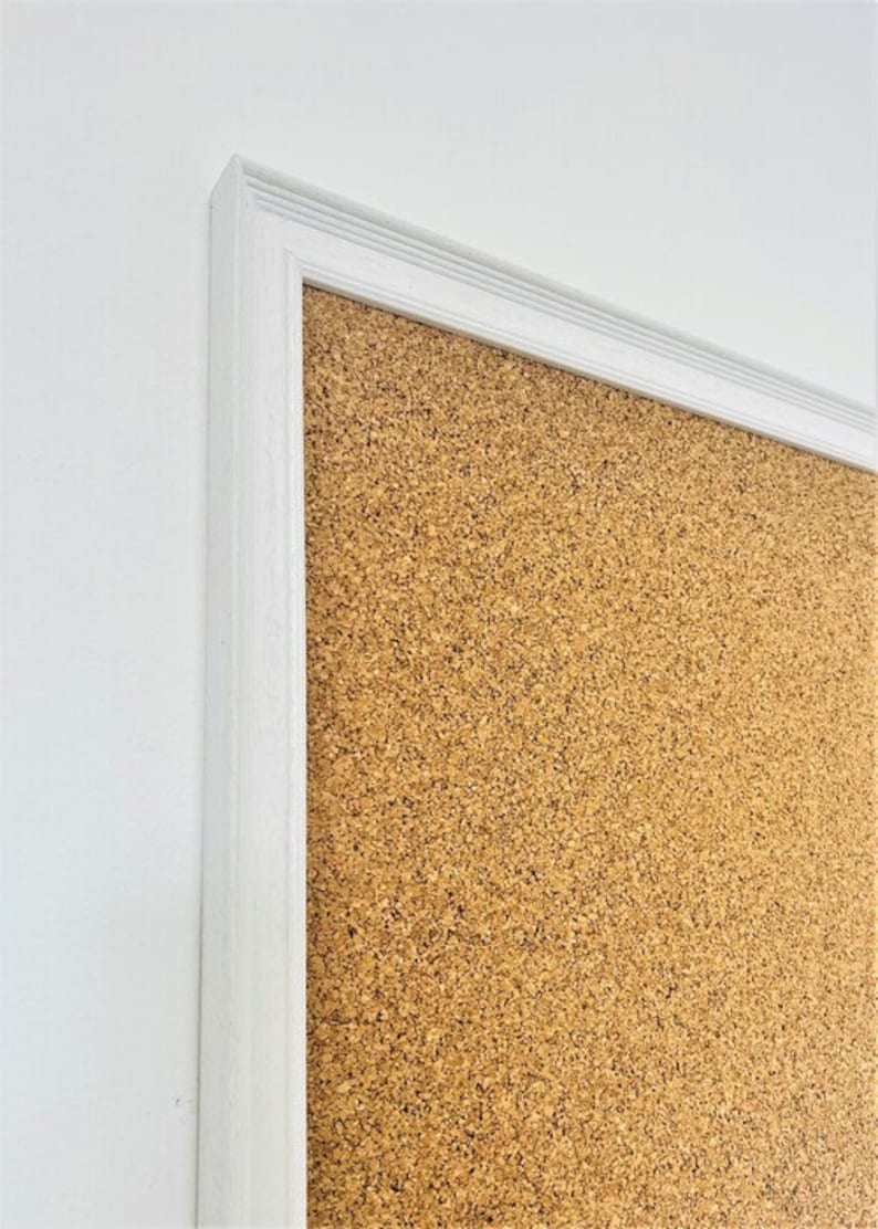 May include: A white framed corkboard with a brown cork surface. The corkboard is mounted on a white wall.
