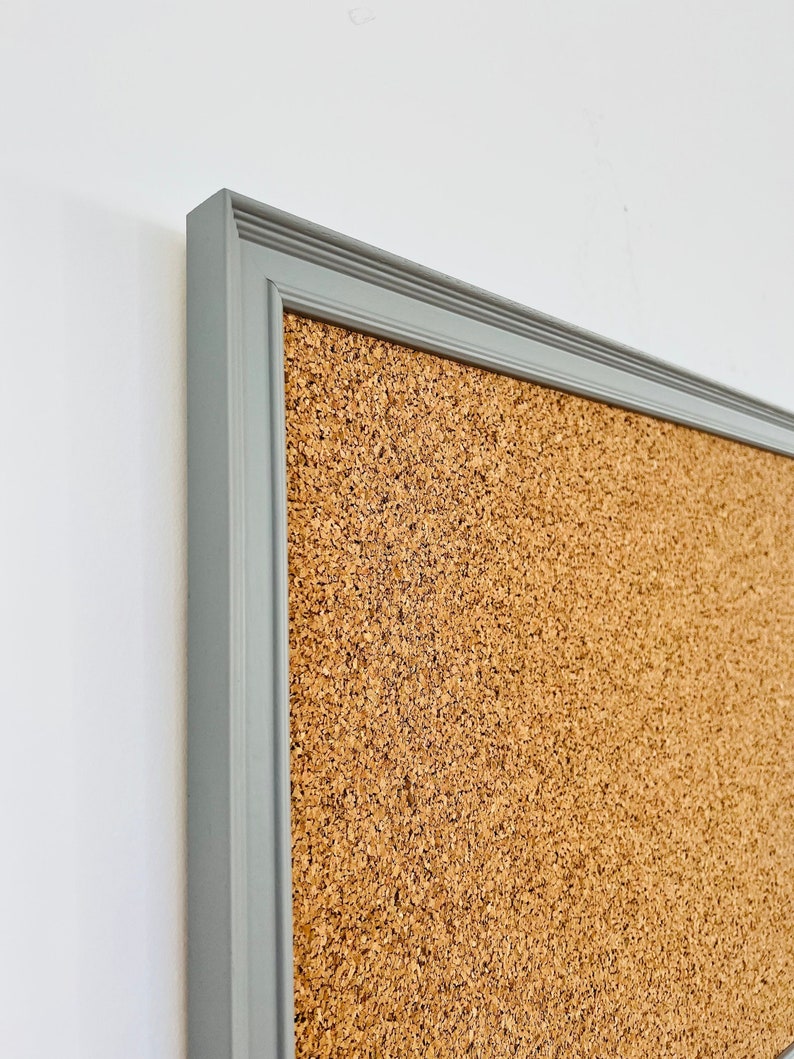 May include: A gray framed corkboard with a textured surface. The frame has a simple, slightly raised molding.