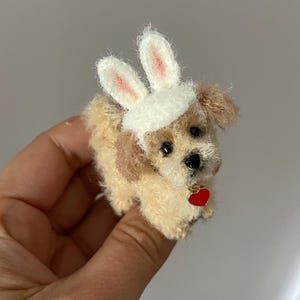 May include: A small, cream-colored, crocheted dog wearing a white bunny hat with pink ears and a red heart charm around its neck.