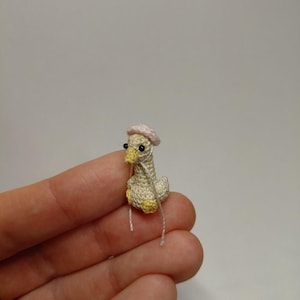 May include: A miniature crocheted duck wearing a light pink beret. The duck is primarily cream-colored with yellow accents on its feet and beak. It has small black eyes and two white strings extending from its body.