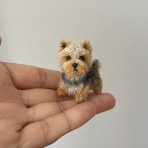 Puede incluir: Una figura en miniatura de un perro Yorkshire Terrier con pelaje texturizado de color tostado y gris oscuro, orejas puntiagudas y una pequeña nariz negra. La diminuta escultura presenta detalles realistas en la cara y el pelaje.
