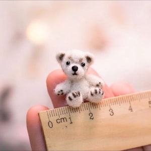 May include: A miniature white felted bear figurine with dark brown accents on the ears, nose, and paws. The bear is positioned next to a wooden ruler, showing a height of approximately 2 centimeters. The bear has small black bead eyes.