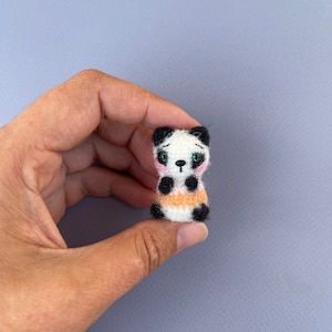 Puede incluir: Una pequeña figura de oso panda hecha a mano. El panda tiene un cuerpo blanco con marcas negras en las orejas, los ojos y las extremidades. Tiene una franja naranja alrededor del centro. La cara del panda tiene mejillas rosadas y una expresión triste.