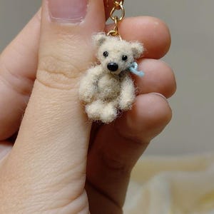 Könnte beinhalten: Ein kleiner, nadelgefilzter Teddybär-Anhänger in Cremefarben, befestigt an einem goldfarbenen Metallring. Der Bär hat kleine schwarze Perlenaugen, eine schwarze Nase und ein hellblaues Band um den Hals, gefertigt mit einer flauschigen Textur.