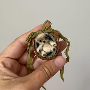 May include: A miniature cream-colored felted bear figurine nestled inside a brown walnut shell. The bear has dark eyes and dark paw pads. A green ribbon is tied in a bow at the top of the walnut shell, with two long ribbon ends hanging down.