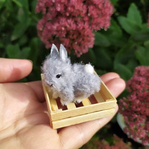 May include: A small gray and white plush bunny rabbit sitting in a wooden crate. The bunny is about 3 inches tall and has a fluffy tail.