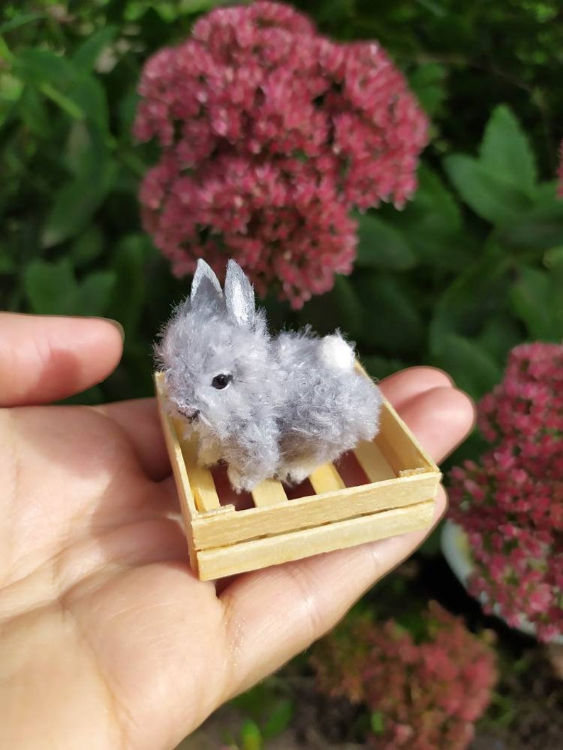 May include: A small gray and white plush bunny rabbit sitting in a wooden crate. The bunny is about 3 inches tall and has a fluffy tail.