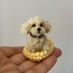May include: A miniature, handmade beige dog figurine with a curly coat, wearing a burgundy collar with a gold bell. The dog sits in a yellow crocheted bed. The figurine is a charming example of handcrafted artistry.