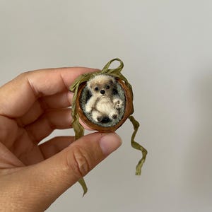 Peut inclure: Une figurine d'ours miniature en feutrine de couleur crème nichée dans une coquille de noix brune. L'ours a des yeux et un nez foncés. Un fin ruban vert olive est noué autour du haut de la coquille de noix, avec deux longs pans pendants.