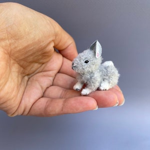 May include: A small gray and white stuffed rabbit with a pink nose and white feet. The rabbit is sitting on a hand.