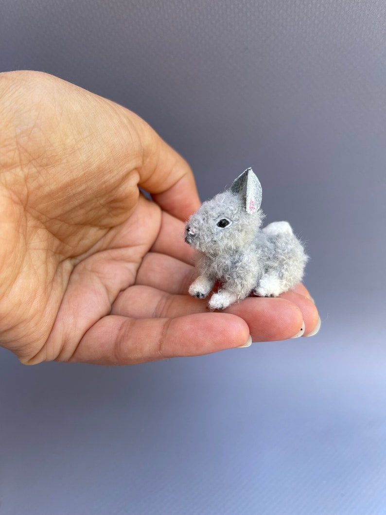 May include: A small gray and white stuffed rabbit with a pink nose and white feet. The rabbit is sitting on a hand.