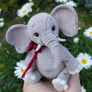 May include: A small, gray, crocheted elephant plush toy with large ears, a trunk, and white tusks. The elephant features black safety eyes and a dark red ribbon tied around its neck. The feet and hands have white crocheted details on the tips.