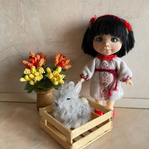 May include: A small doll wearing a white embroidered dress with red accents and a red headband with flowers stands next to a wooden crate with a gray fluffy bunny inside.