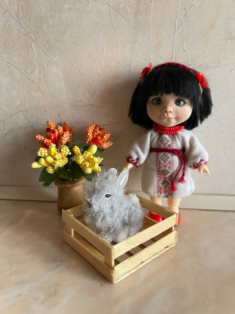 May include: A small doll wearing a white embroidered dress with red accents and a red headband with flowers stands next to a wooden crate with a gray fluffy bunny inside.