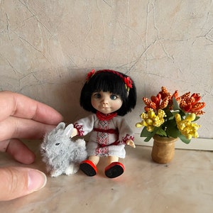 May include: A small doll wearing a white embroidered dress with red accents and a red flower crown. The doll has black hair and blue eyes. The doll is sitting next to a gray plush bunny rabbit. A small potted plant with yellow and orange flowers is in the background.