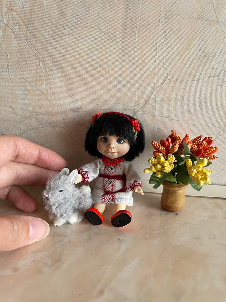 May include: A small doll wearing a white embroidered dress with red accents and a red flower crown. The doll has black hair and blue eyes. The doll is sitting next to a gray plush bunny rabbit. A small potted plant with yellow and orange flowers is in the background.