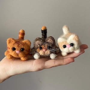 Op de afbeelding: Drie vilten wollen kattenfiguren. De kat links is oranje en wit, de middelste kat is bruin en wit en de kat rechts is wit met oranje markeringen. Alle drie katten hebben zwarte ogen en roze neuzen.