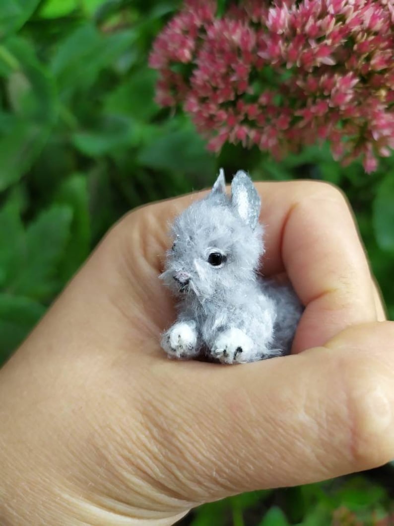May include: A small, gray, plush bunny rabbit with white feet and black eyes. The bunny is sitting on a hand.