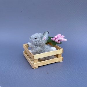 May include: A small gray bunny rabbit sits in a wooden crate with a pink flower.